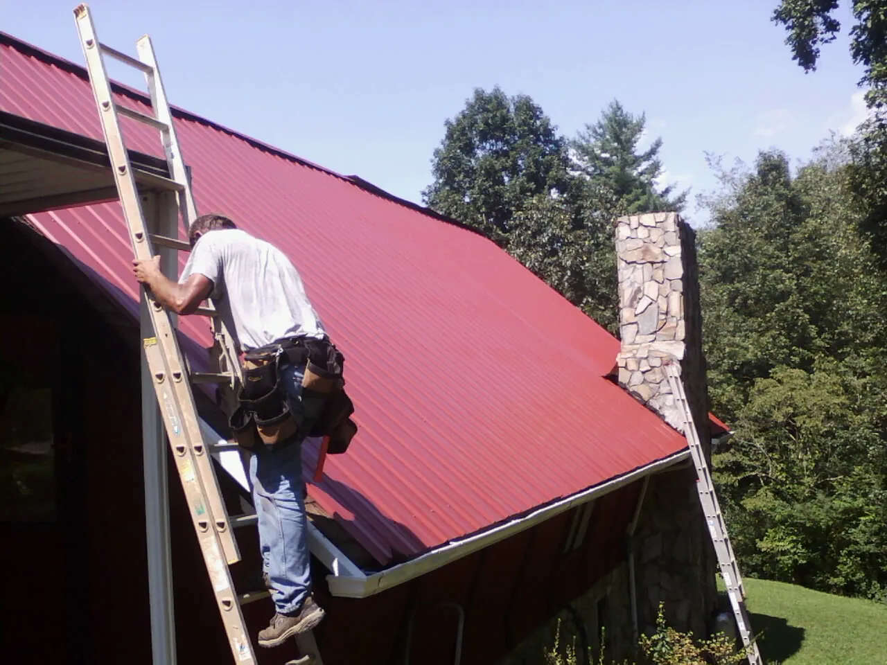 Roofer on a ladder beside a red metal roof and stone chimney — Soffit & Fascia Repair by Roofing Brothers in Mccleary